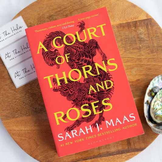 An image of the book "A Court of Thorns and Roses" on a wooden board with paua shells and branded stickers framing it. - Second-Hand books from At The Helm
