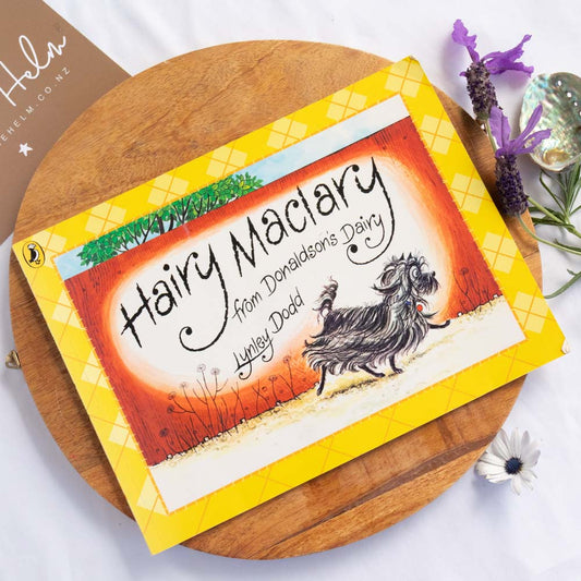 A yellow and orange children's book titled 'Hairy Maclary from Donaldson's Dairy' by Lynley Dodd, styled on a round wooden board with decorative flowers and a small card in the background.